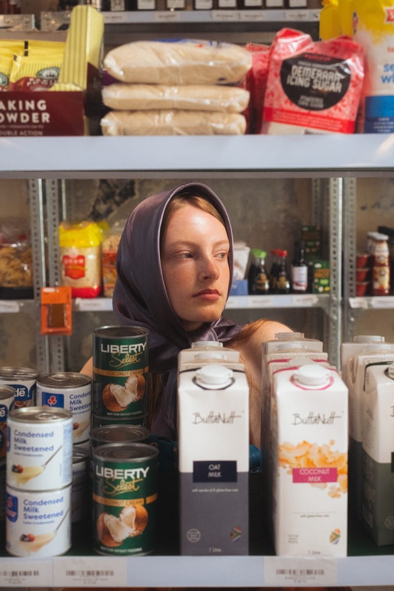 Model standing behind a shelf of tinned milk and cartons, framed by the shop stock