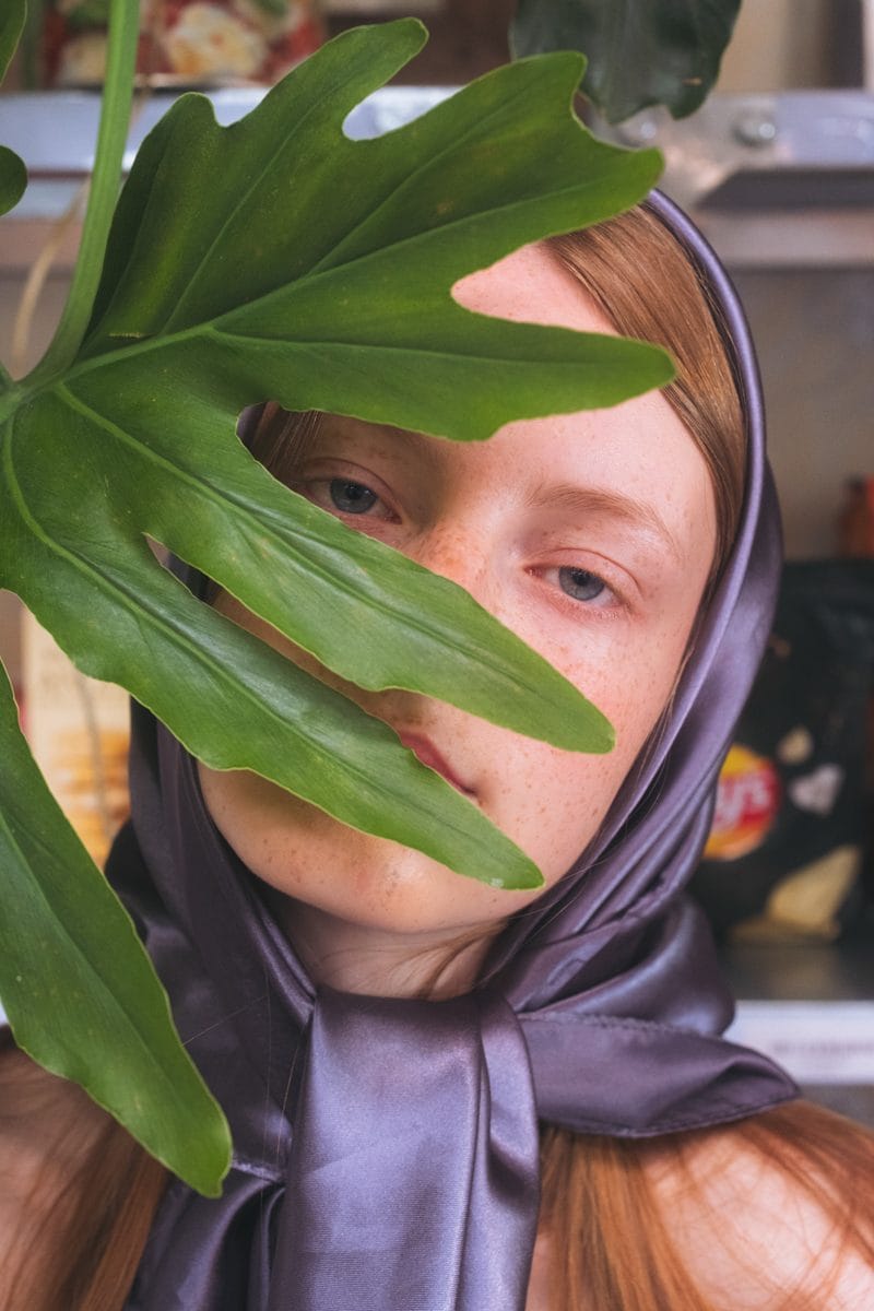 Close portrait, a philodendron leaf held across the face over a lilac silk headscarf