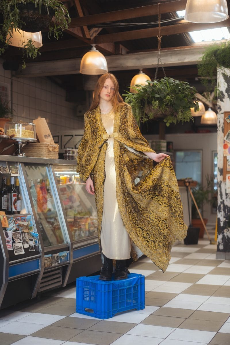 Model standing on a crate in the deli, gold brocade coat by Viviers open over a cream slip dress