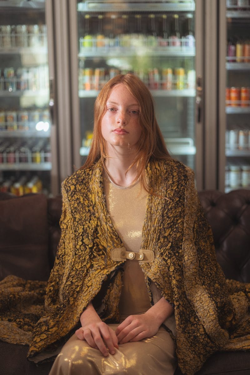 Seated portrait in front of the drinks fridges, gold brocade coat and cream slip dress