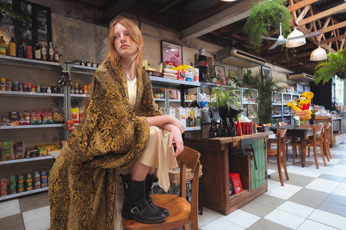 Model seated on the counter of the general store, gold brocade coat and black boots, aisles receding behind her