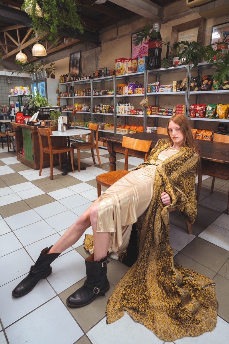 Model seated at a deli table, the gold brocade coat spread across the chequered floor