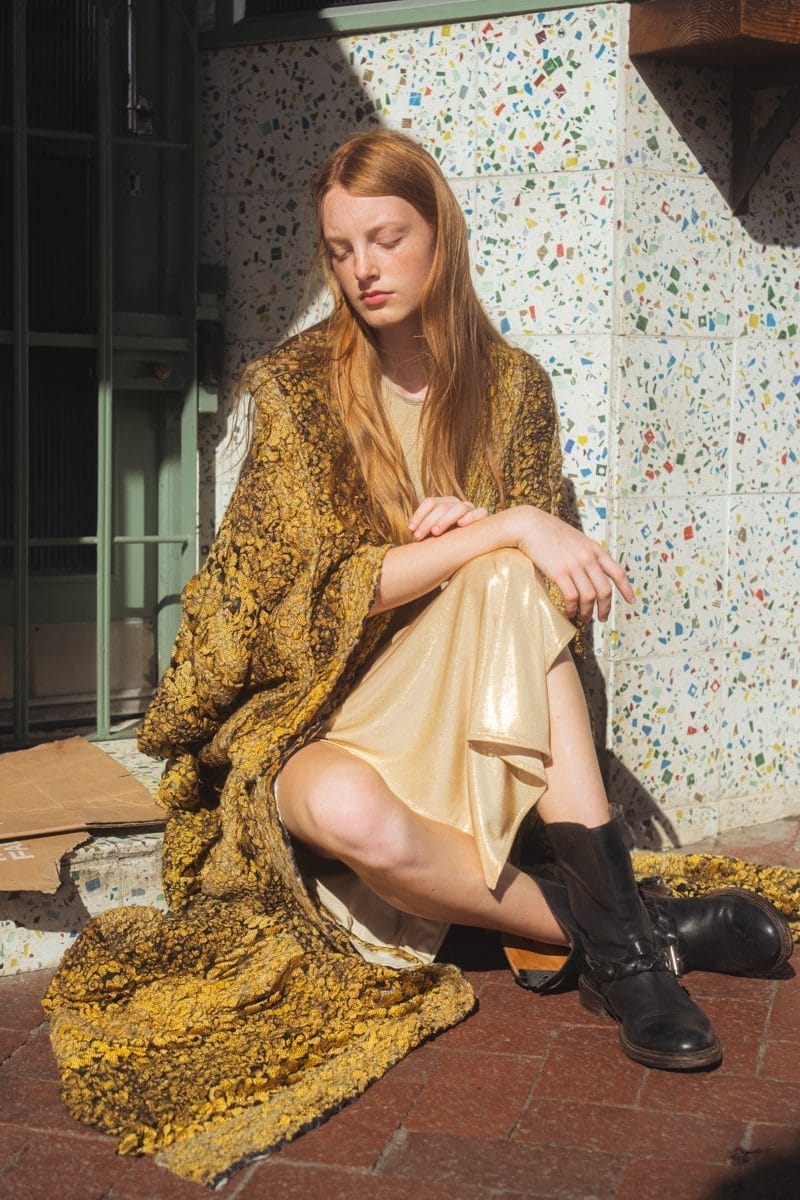 Model seated outside against a speckled wall, gold brocade coat by Viviers and black boots
