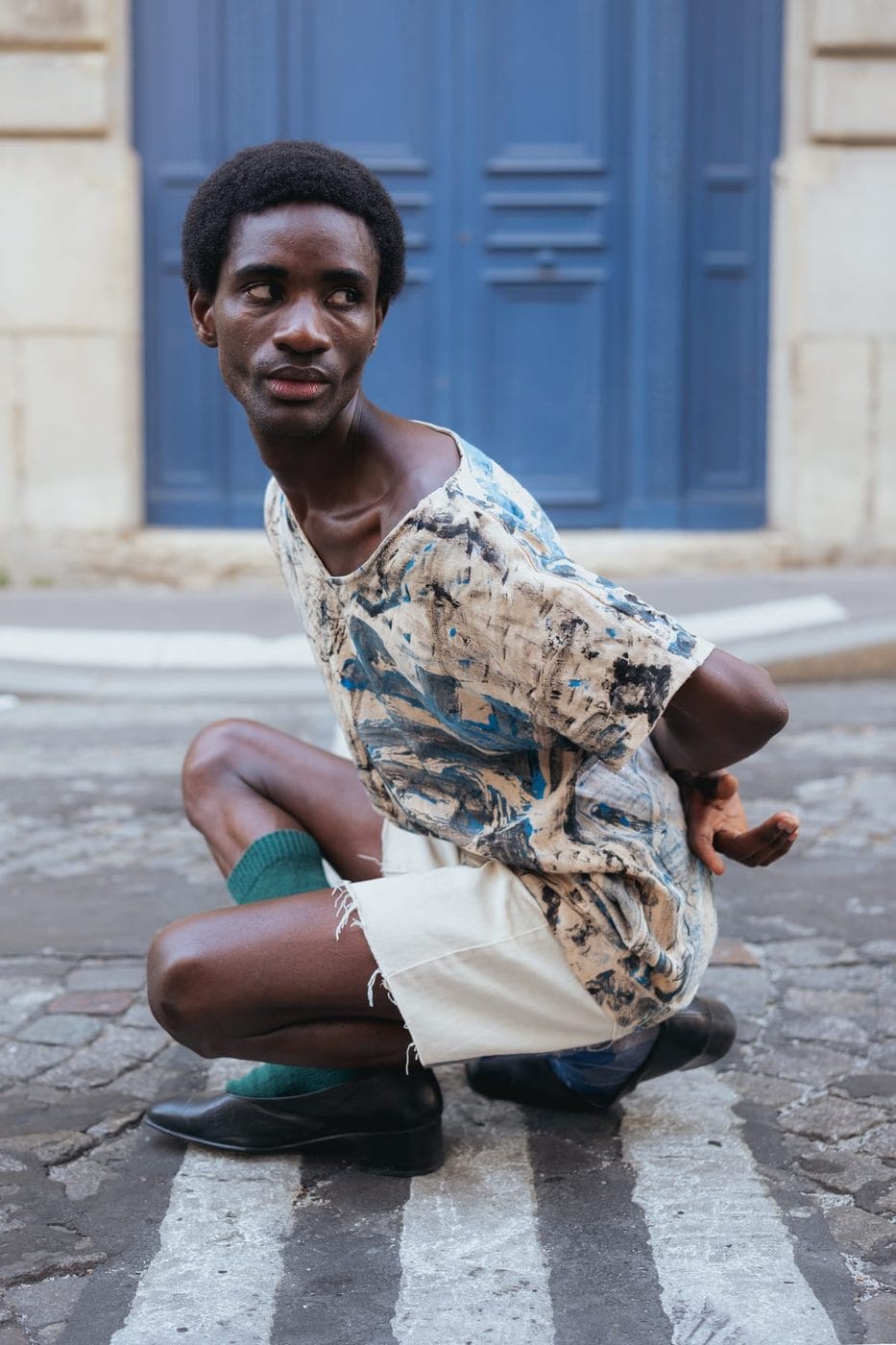 Homme accroupi en chemise à motifs devant une porte bleue, @modelmodu
