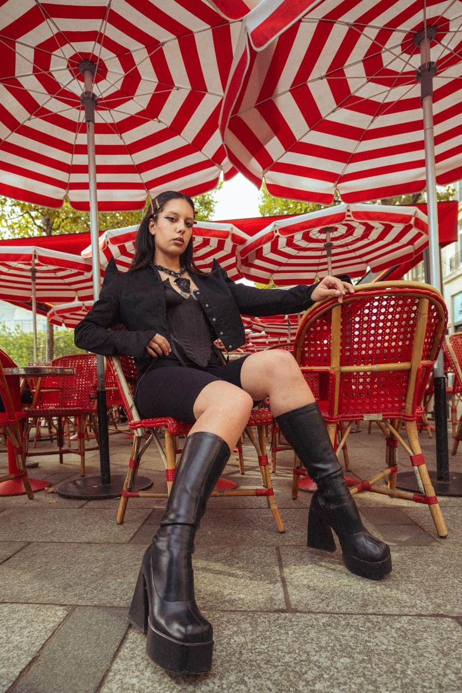 Femme en noir assise sous des parasols de café rayés rouge et blanc, bottes à plateforme, June Viktoria Anderson (@junyeanderson)