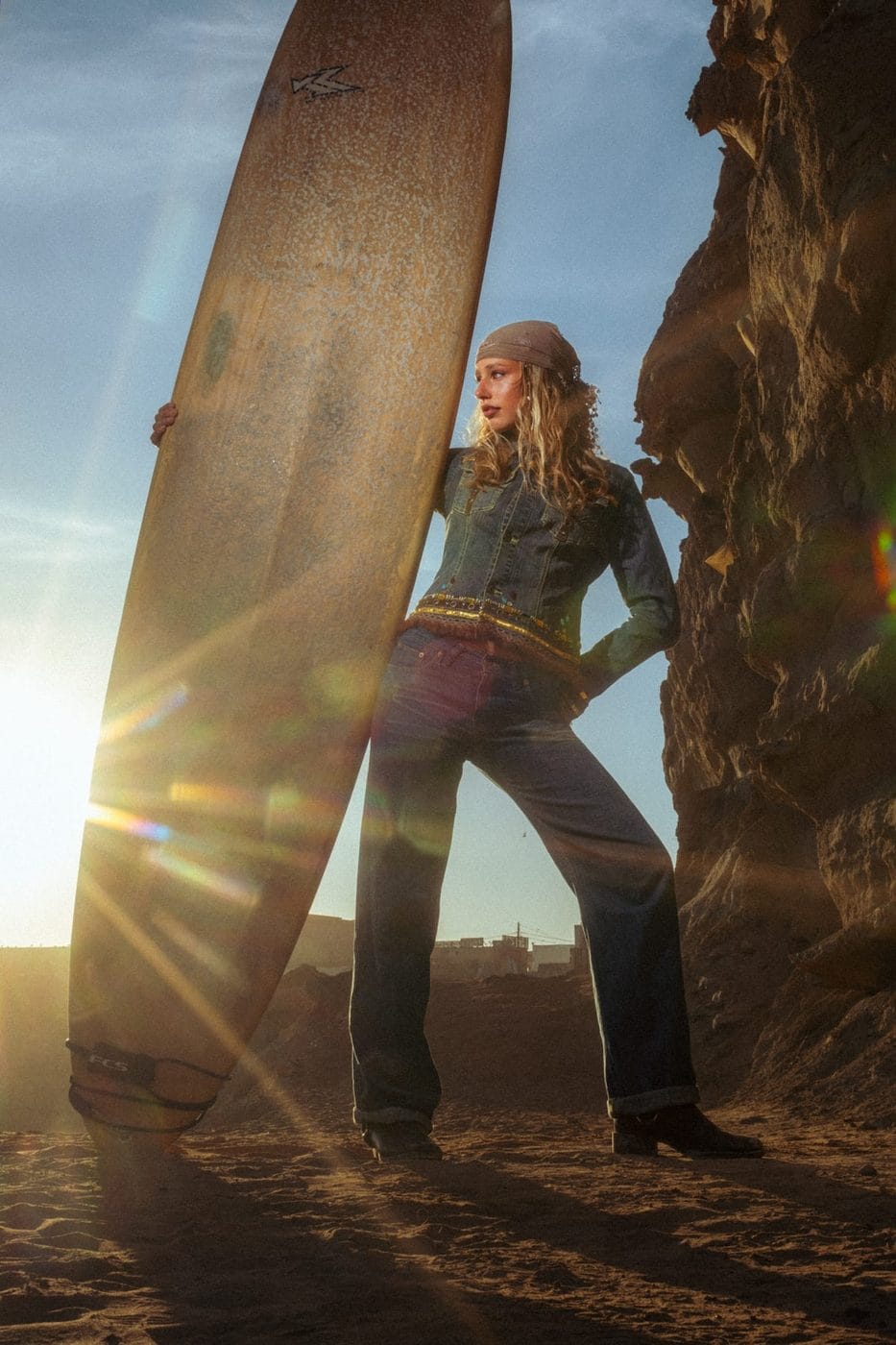 Femme en denim adossée à une planche de surf au coucher du soleil, à contre-jour, Merle (@merlecrohn)