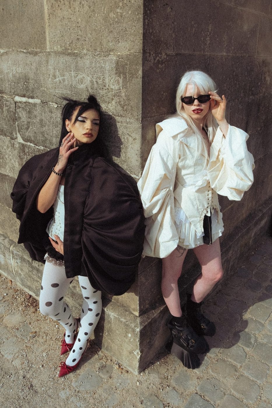 Deux femmes contre un mur de pierre, l'une en cape noire et collants à pois, l'autre en blanc, Gabriella Magniez (@gxbxxy) et Victoria Laurent (@2moiselle.savage), Stylisme asgoodasgold.mt, maquillage annabeth.mua