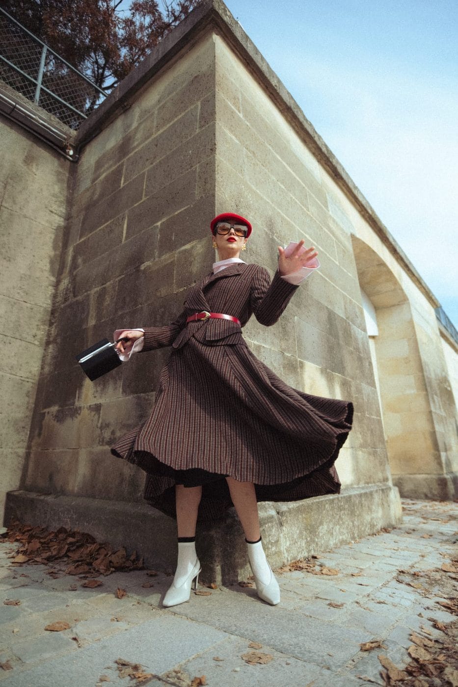 Femme en robe plissée brune et béret rouge tournoyant le long d'un mur de pierre, @olga_kuzymko