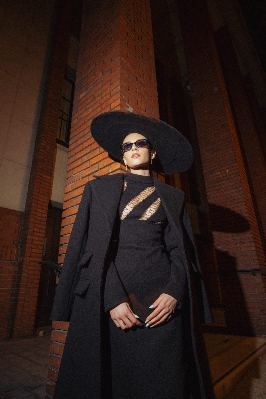 Femme en long manteau noir et chapeau à large bord devant un mur de brique, la nuit, Amélie Diaz, Ses propres créations